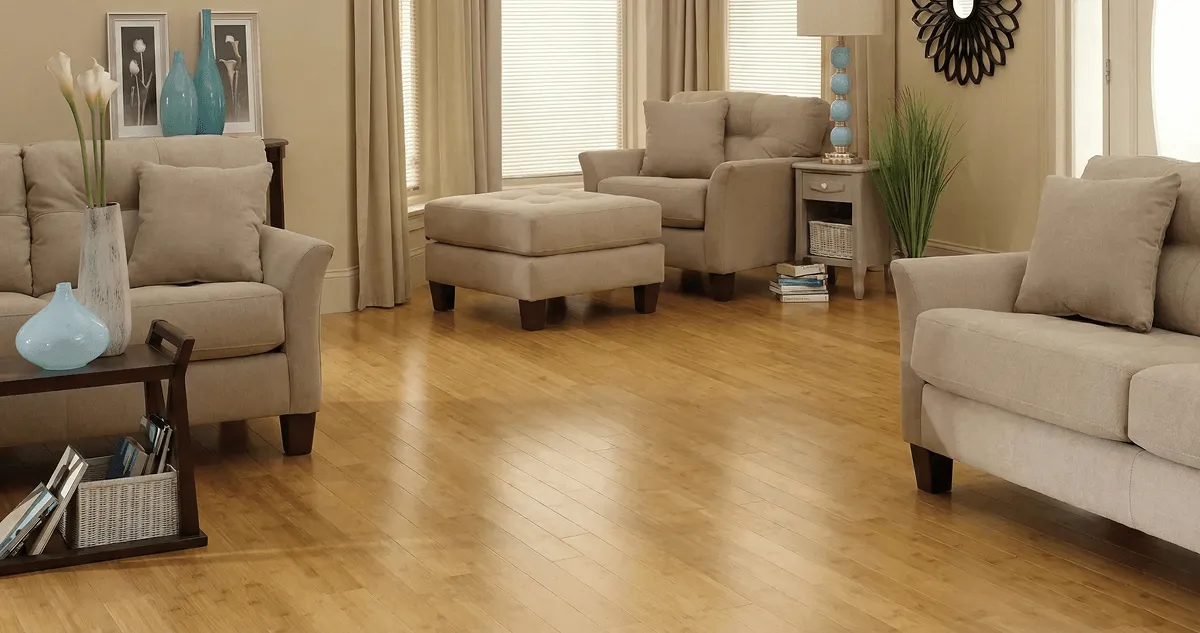A living room with professionally installed bamboo flooring, beige sofas, a matching ottoman, a side table with a blue lamp, vases, framed pictures, and large windows with blinds and curtains in Indianapolis, IN
